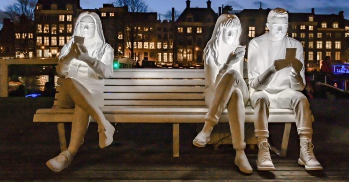 White mannequins illuminated by the screen of their own smartphone, sitting on a street bench