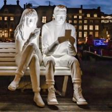 White mannequins illuminated by the screen of their own smartphone, sitting on a street bench